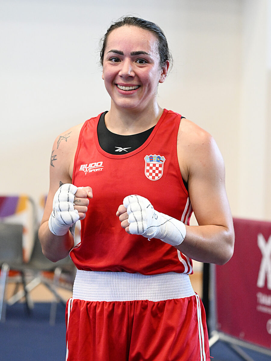A smiling athlete in a red boxing uniform with Croatian insignia and white gloves stands ready, fists clenched.