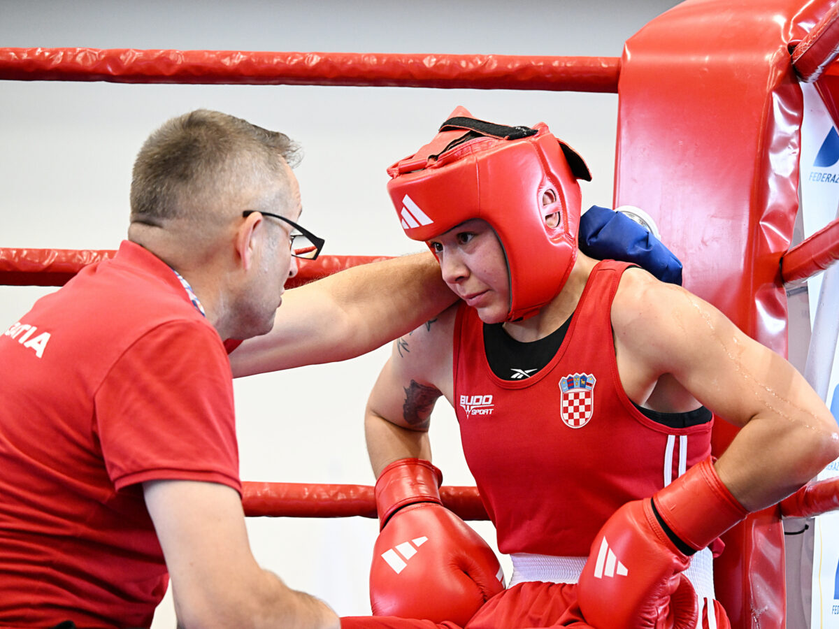A boxer in red gear receives encouragement from a coach inside a boxing ring.