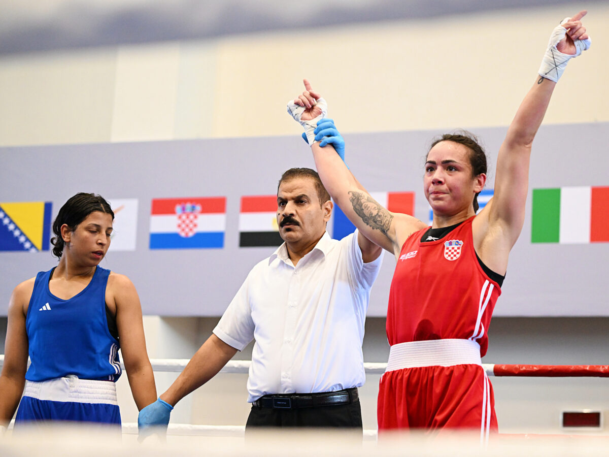A referee raises the arm of a boxer in a red uniform to declare victory, while another boxer in blue stands beside them, with flags of various nations displayed in the background.