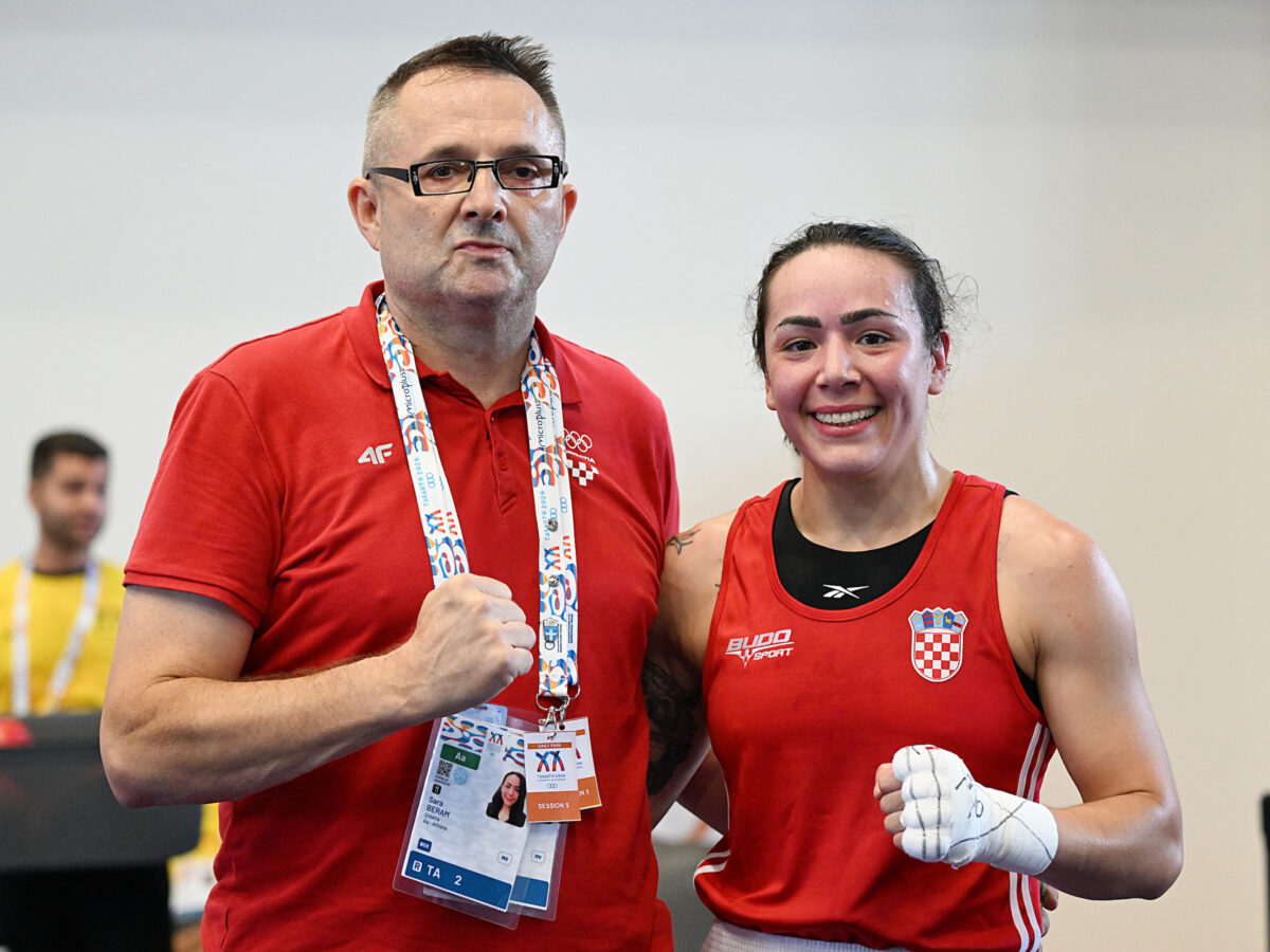 A person in a red polo shirt with event lanyards stands beside a boxer in a red athletic top, both making fist gestures.