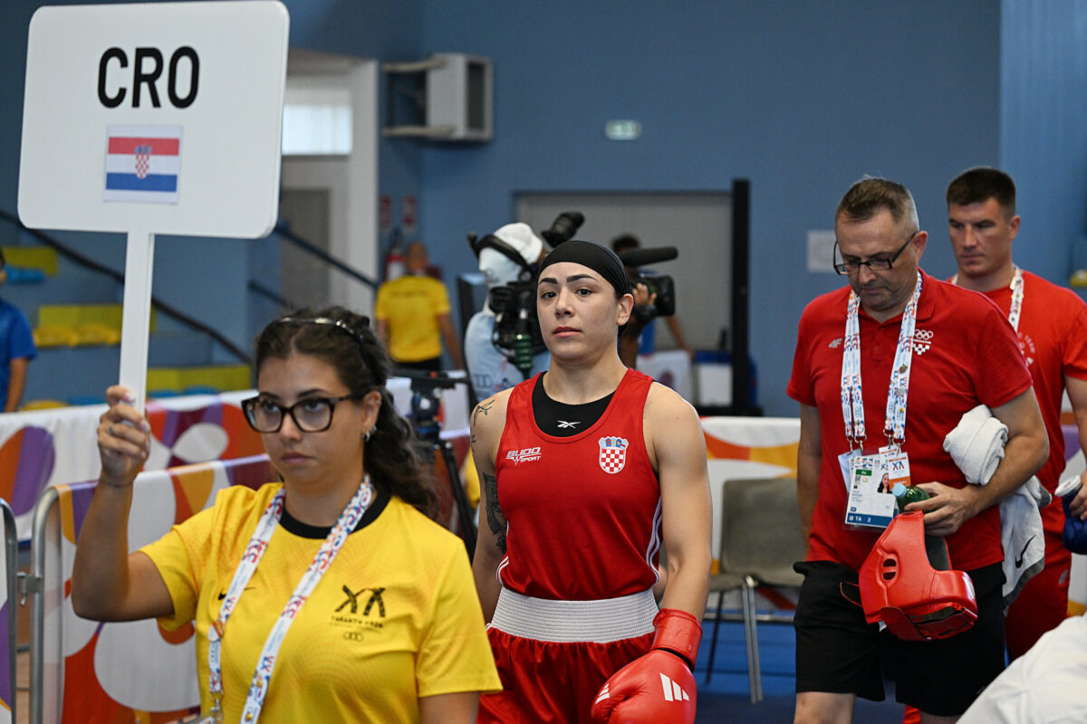 A boxer in a red uniform walks alongside team staff at a sports event, with a volunteer holding a sign indicating Croatia.